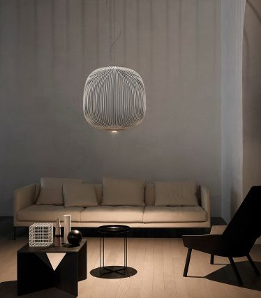 Ett minimalistiskt vardagsrum med pendellampan Spokes från Foscarini som elegant blickfång. Lampans filigrana metallstänger kastar mjuka ljusmönster på vägg och tak, vilket ger rummet en lätt, skulptural effekt. En ljus soffa, mörka accenter och naturmaterial skapar en lugn, modern atmosfär