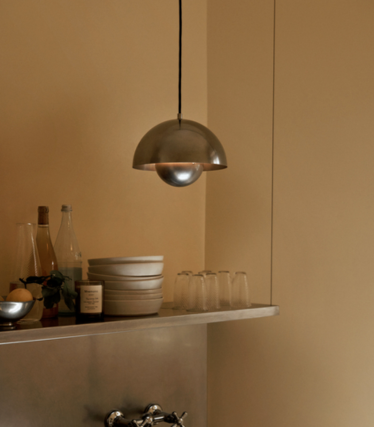Pendellampa i polerad metall över en hylla med keramiska skålar och glas framför en sandfärgad vägg.