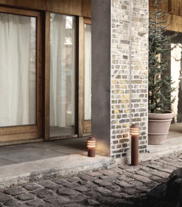 Utomhusscen med två moderna cylindriska gångbelysningsarmaturer placerade bredvid en stenbelagd gångväg och en pelare av tegel och betong. Bakom dem finns glasdörrar med träramar, ljusridåer och en stor krukväxt. Det varma skenet från belysningen skapar en mysig och inbjudande atmosfär.