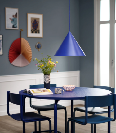 Blå konformad pendellampa över ett runt blått matbord med en blombukett, i en matsal med blå vägg och konstverk.