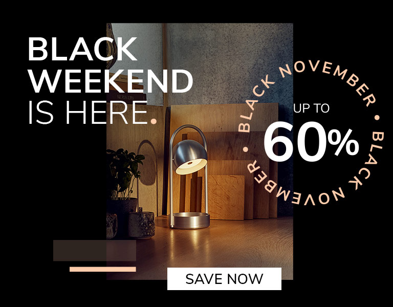 Modern silver table lamp glowing on a wooden desk with cutting boards and plants in the background. Promotional text reads: ‘Black Weekend is here. Up to 60%. Black November.’ A button says ‘Save Now.